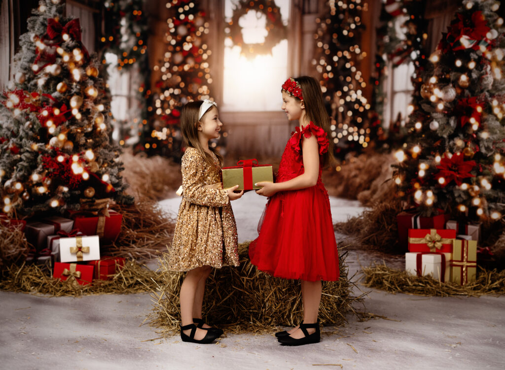 Atlanta holiday mini session of two girls in red and gold dresses giving a present to each other. backdrop is a Christmas decorated barn with lighted trees.