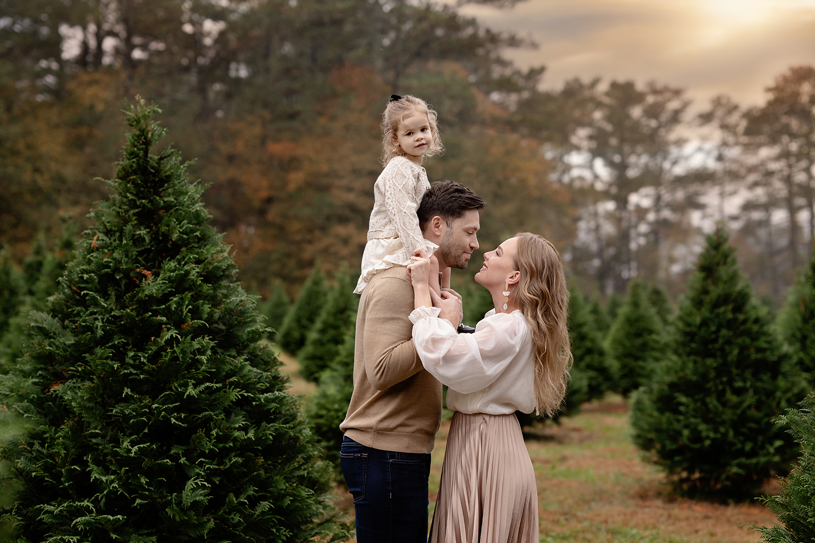 Toddler girl on Dad's shoulders and Mom looking lovingly at Dad. Photographed by Atlanta Family photographer on a tree farm.