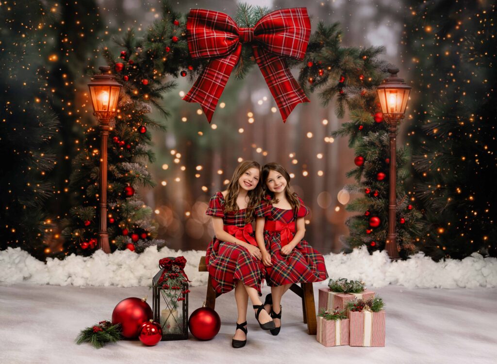 Two girls in plaid dresses sitting on a bench smiling in front of a Christmas arch backdrop. 