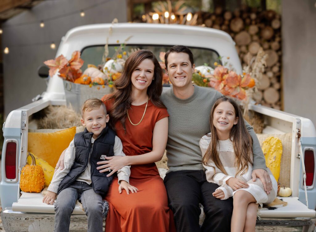 Family sitting on truck bed that is decorated with fall decor and smiling at camera.