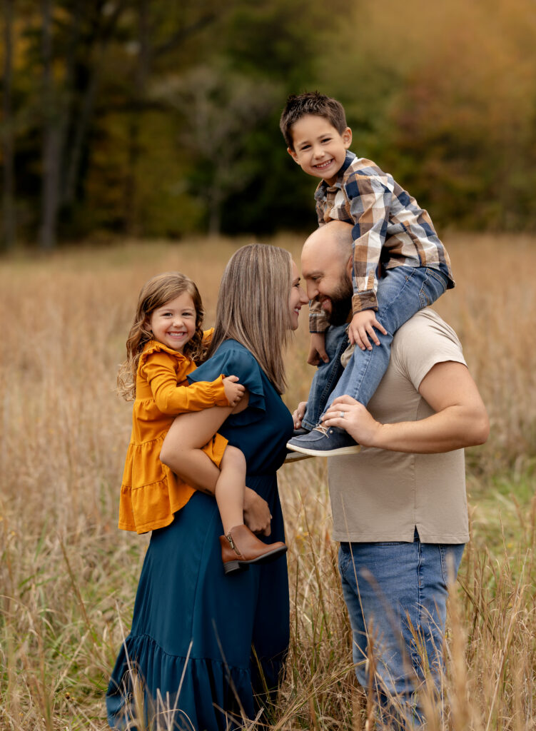 Family of 4 in hay field. Boy on Dad's shoulders and little girl on Mom's back smiling at the camera.