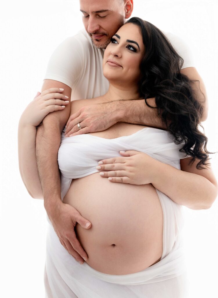 Pregnant women in white drape with husband hugging her from behind.