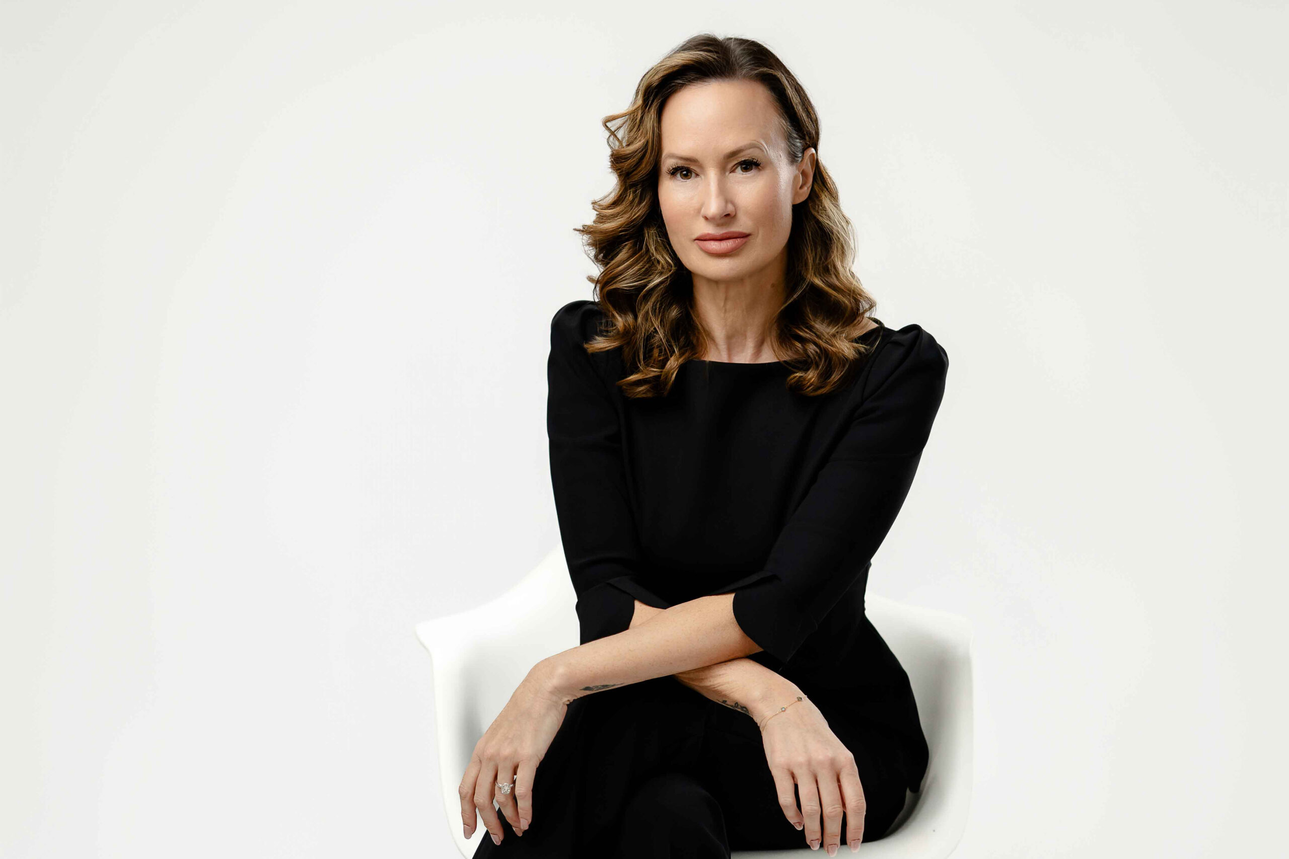 Women in black suit, sitting in a white chair with arms on knee and having a more serious expression for her professional business photo in Atlanta.