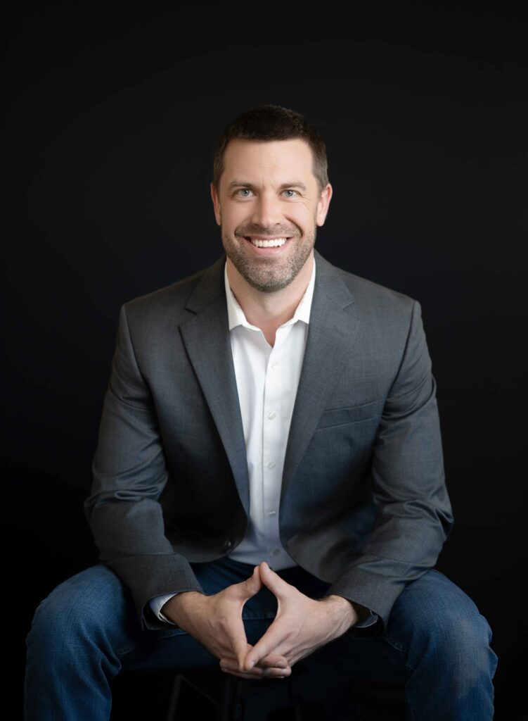Man with gray sports coat on, sitting on stool and taking professional business photo.