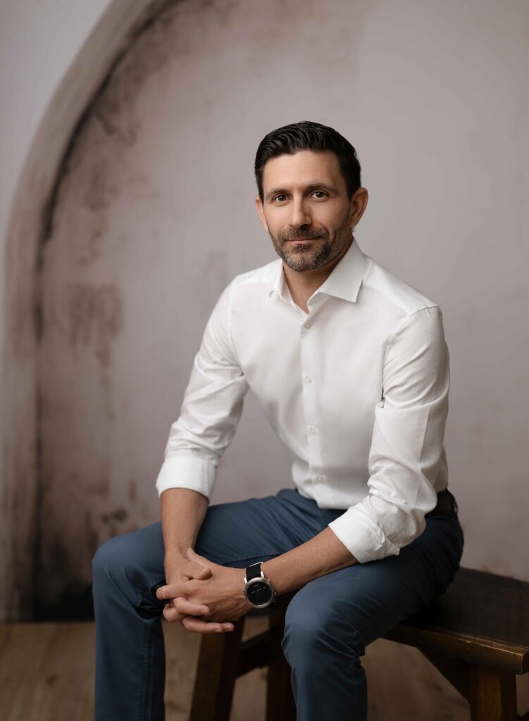 Man sitting on stool in front of textured arch wall, taking a business photo.