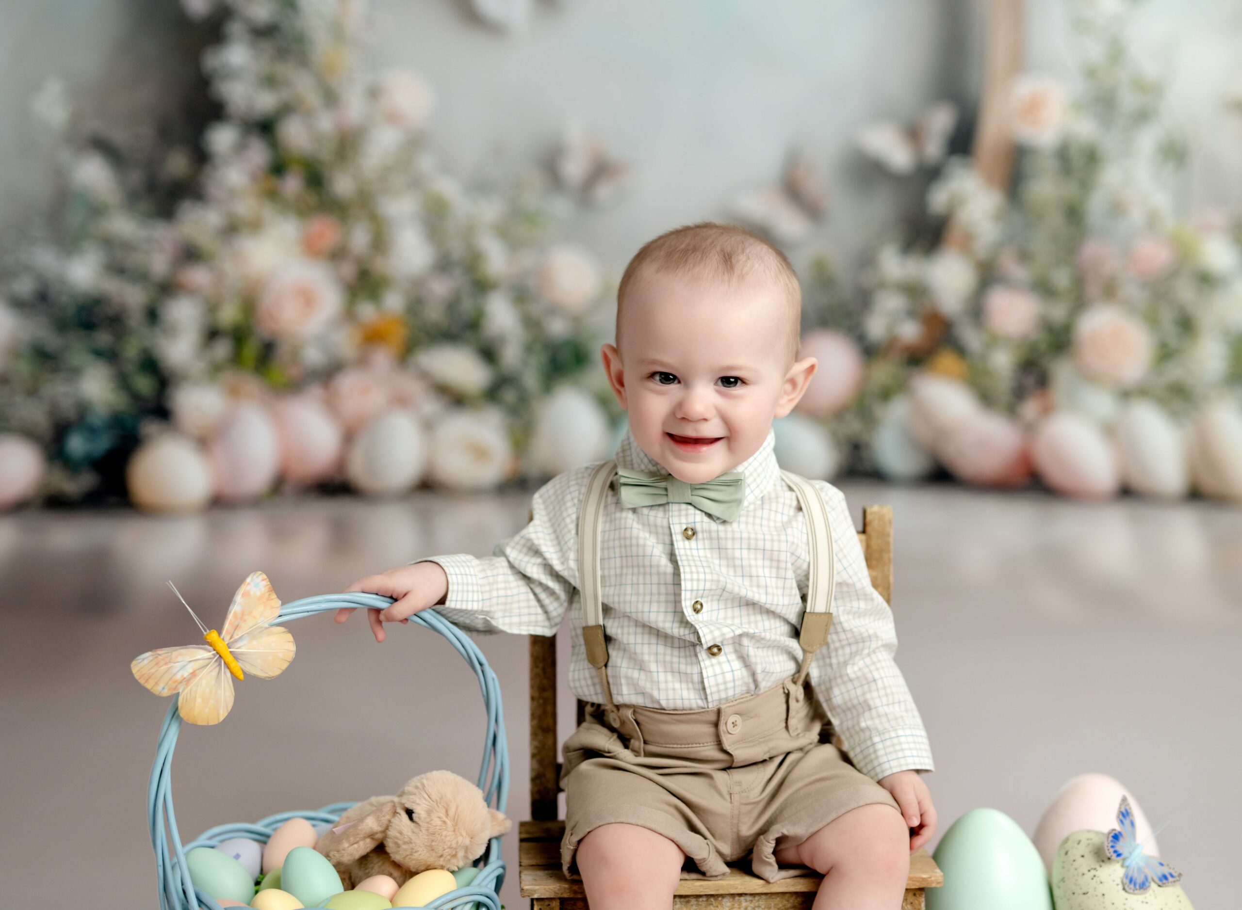 Smiling baby boy sitting on a wooden chair with Easter basket and pastel eggs during an Atlanta baby photography Easter mini session in a luxury studio setting.